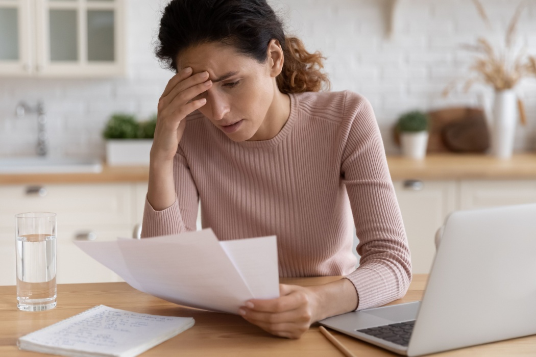 Stressed young woman looking at paperwork while using laptop.