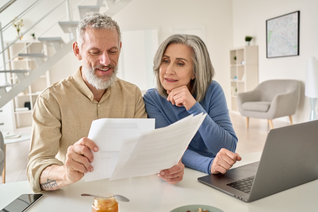 A senior couple sit at a laptop comparing multiple documents