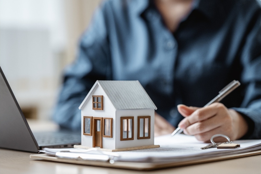A person signing a document with a model house on the table.